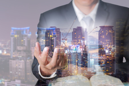 Double exposure of businessman hand hold, construction crane, building, city and urban in the twilight or night as industrial and business concept.の写真素材