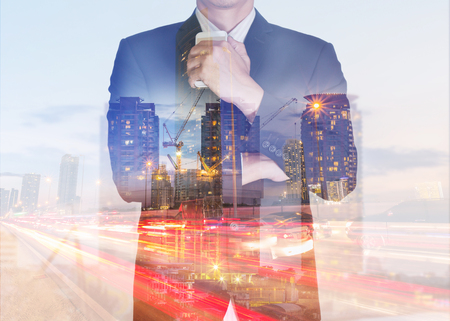 Double exposure of businessman stand up and think idea about business, light trails city, urban street, construction crane and building in the evening, twilight as thoughtful concept.の写真素材