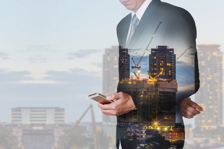 Double exposure of businessman stand up and research by smartphone, construction crane and building in the evening, twilight as technology, industrial and business concept.の写真素材