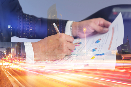 Double exposure of businessman hand hold a pen write, review document, light trails on the street and urban at dusk, night as business, review and working Concept.の写真素材