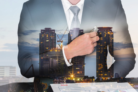 Double exposure of businessman holding a pen on the pocket, construction crane and building in the evening, twilight as business, analysis and industrial conceptの写真素材
