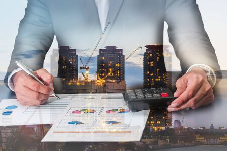 Double exposure of businessman working with document and calculator for analyze in the work, construction crane and building in the evening, twilight as analysis and commitment concept.の写真素材