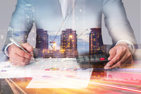 Double exposure of businessman working with document and calculator analyze, construction crane, light trails street, urban and building in the evening, twilight as analysis and commitment concept.の写真素材