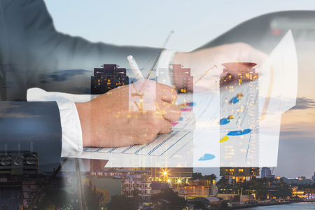 Double exposure of businessman hand hold a pen write, review document, construction crane and building in the evening, twilight as industry, business, review and working Concept.の写真素材