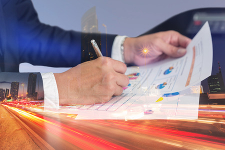 Double exposure of businessman hand hold a pen write, review document, light trails on the street and urban at dusk, night as business, review and working Concept.の写真素材