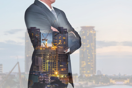 Double exposure of Businessman arms crossed and stand up, construction crane and building in the evening, twilight as business, thinking vision of leader and construction concept.の写真素材