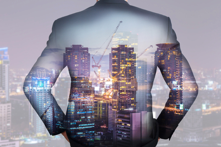 A back turned businessman, construction crane, building, city and urban in the twilight or night as business, vision of leader and industrial conceptの写真素材