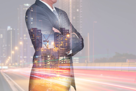 Double exposure of Businessman arms crossed and stand up, light trails on the street urban and construction crane and building in the night as business, thinking vision of leader and industry concept.の写真素材