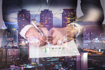Double exposure of businessman hand hold a pen point to the bar graph, construction crane, building, city and urban in the twilight or night as Business, commitment and analysis conceptの写真素材