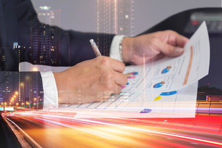 Double exposure of businessman hand hold a pen write, review document, light trails on the street and urban in the night as business, review and working Concept.の写真素材