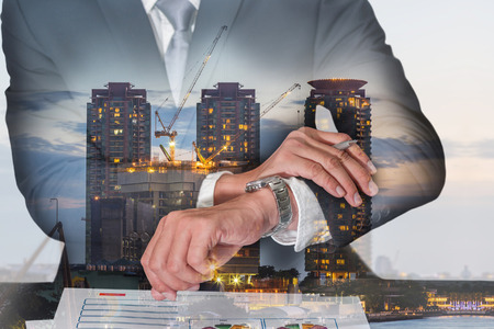Double exposure of businessman look the watch, construction crane and building in the evening, twilight as appointment, business and industrial concept.の写真素材