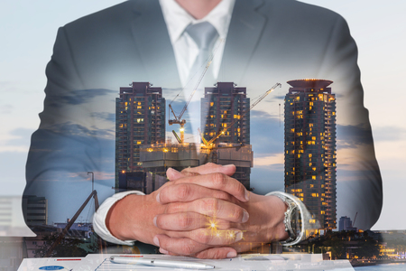 Double exposure of businessman fingers crossed and sit down, construction crane and building in the evening, twilight as meeting, commitment, business and industrial concept.の写真素材