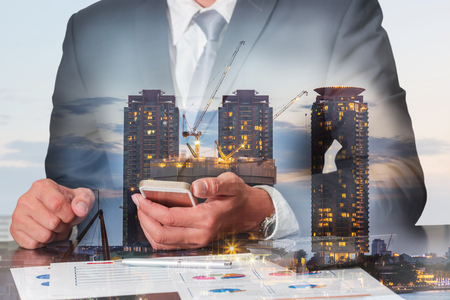 Double exposure of businessman working with smartphone construction crane and building in the evening, twilight as commitment, technology, communication and industrial concept.の写真素材