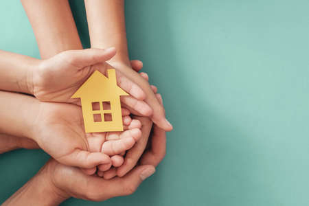 Hands holding yellow paper house on blue background, family home, homeless shelter housing and home...