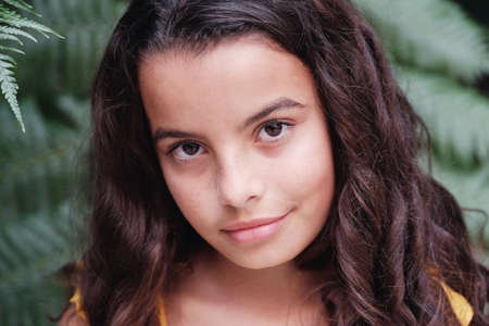 Close up portrait of a confident, charming and gorgeous mixed multicultural preteen girl with beautiful curly hair over grean tropical leaves, looking at cameraの写真素材