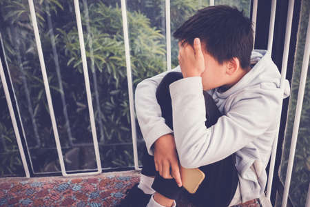 Young Asian preteen teenager boy hugging his knee and cover his face and holding a smartphone, Cyber bullying in kid, depressed child mental healthの写真素材
