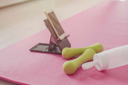 Dumbbells, water bottles and smartphone on pink floor exercise mat, workout training at home, online streaming videos excercise, social distancing conceptの写真素材