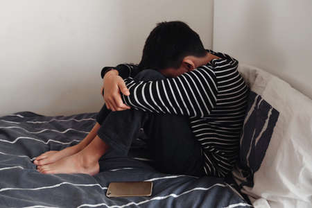 Young Asian preteen teenager boy hugging his knee in his bedroom with smartphone, Cyber bullying in kid, depressed child mental healthの写真素材