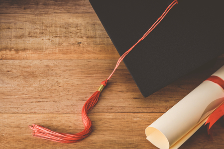 mortarboard or graduation cap with red tassel and diploma tied with red ribbon on wooden table, vintage toneの写真素材