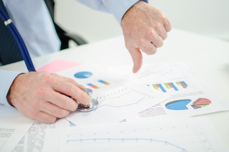 Businessman before financial graphs with a stethoscope, thumb downの写真素材