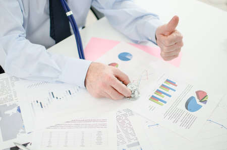 Businessman before financial graphs with a stethoscope, thumb upの写真素材