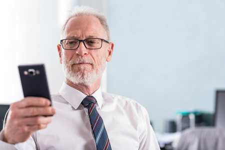 Portrait of senior businessman looking at his mobile phone at officeの写真素材