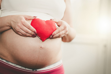 Pregnant woman holding a red heart in front of her bellyの写真素材