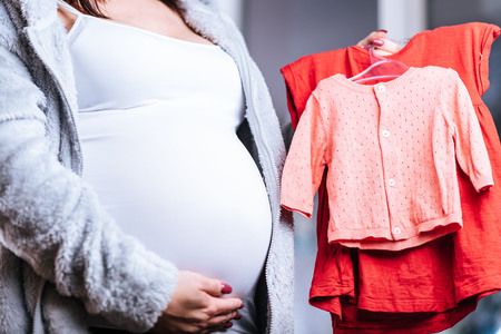 Pregnant woman holding baby clothes and touching her bellyの写真素材