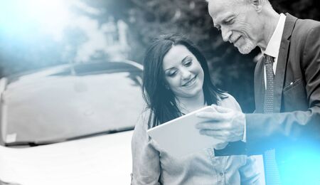 Car salesman giving informations on tablet to young woman outdoors, light effectの写真素材