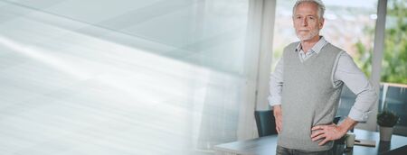 Portrait of senior businessman standing in office with hands on hips; panoramic banner