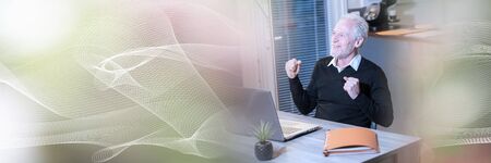 Smiling senior businessman showing success gesture in front of his laptop; panoramic banner