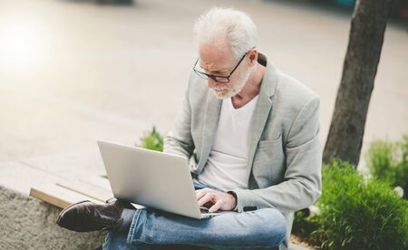 Mature businessman sitting on a bench outdoors and using laptop の写真素材