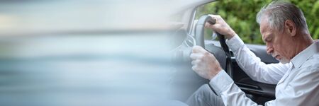 Tired senior man falling asleep behind steering wheel of car; panoramic banner