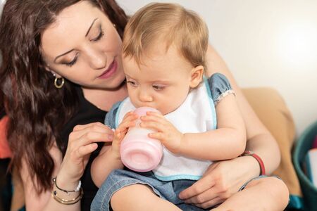 Cute baby girl sitting on her mother and drinking milk from bottleの写真素材