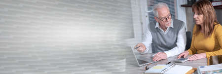 Two mature coworkers working on laptop at office; panoramic banner