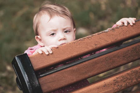 Baby girl playing hide and seek behind a bench, outsideの写真素材