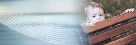 Baby girl playing hide and seek behind a bench, outside; panoramic banner