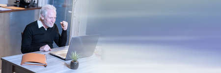 Smiling senior businessman showing success gesture in front of his laptop; panoramic bannerの写真素材