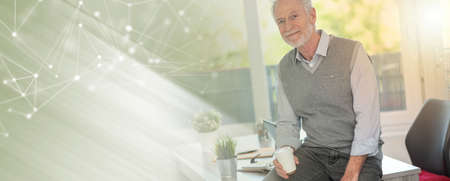 Portrait of smiling senior businessman sitting on his desk in office; panoramic banner
