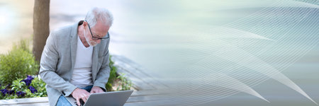 Mature businessman sitting on a bench outdoors and using laptop; panoramic bannerの写真素材