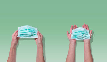 Hands holding face mask for virus preventive measure on light green background; top view
