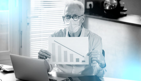 Senior businessman wearing a medical face mask and showing a financial graph with arrow down trend;...