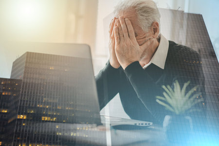 Stressed senior businessman sitting in office with head in hands; multiple exposureの写真素材