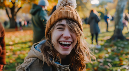 Smiling young woman in a warm hat, laughing joyfully amidst autumn leaves and a park settingの素材
