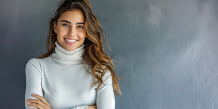 Cheerful young woman with long brown hair in a white sweater, standing confidently indoorsの素材