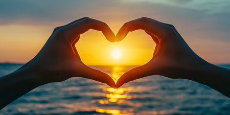 Two hands create a heart shape while a sunset reflects on the ocean waterの素材