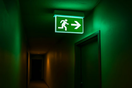 A glowing green emergency exit sign directing towards a right exit in a dim corridorの素材