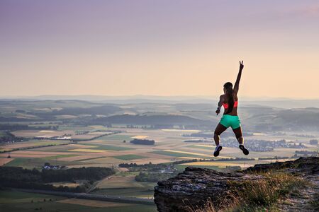 young woman jumping on a rock in front of rural landscape and sunset
