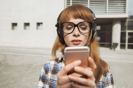 close up of a young hipster woman with red hair that is wearing glasses and uses an application in her smart phone device to send a text messageの写真素材