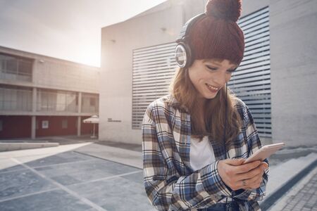 a young woman wearting a beanie and headphones, that is using a music application in her smart phone device while in an urban, modern city architecture environmentの写真素材